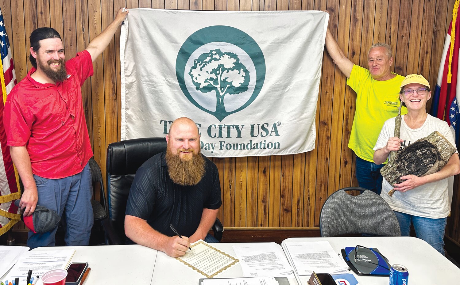 Mayor Jeremy Dawson (seated) signs an Arbor Day proclamation as Parks board member Matthew Klusmeyer and public works director Danny Dougherty hold up the city’s new Tree City USA flag. Alderwoman Bekki Galloway holds piece of tree that was cut after it became damaged by rubbing against a power line.