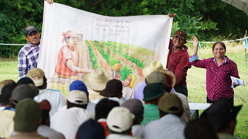 The CIW&rsquo;s Nely Rodriguez (right) leads a conversation on protection against retaliation under the Fair Food Program during a recent Worker-to-Worker Education session on a Fair Food Program Participating Farm, as two volunteers display an FFP drawing depicting a supervisor scolding a worker for speaking up on the job.